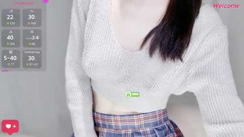 Video of a fair-skinned woman with straight, shoulder-length dark brown hair, wearing a light grey, long-sleeved, V-neck knit sweater and a plaid skirt. Background includes temperature and humidity readouts, with \"Welcome\" text.