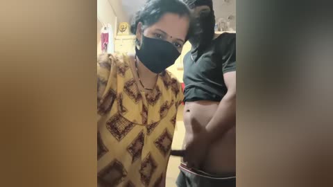 A video shows a pregnant woman with medium skin tone, wearing a yellow patterned shirt, black face mask, and a green shirt lifted, revealing her abdomen. She stands in a cluttered, dimly lit room with a blurred background.