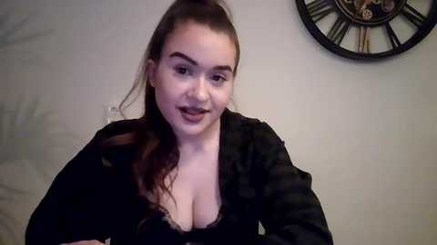 Video of a fair-skinned, young woman with long, wavy brown hair, wearing a black, low-cut top revealing ample cleavage. She has a slight smile and is indoors against a plain wall with a large, decorative clock.