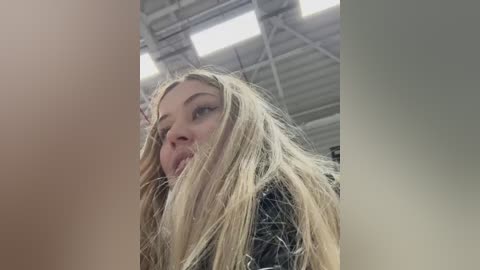 A video of a blonde woman with long hair partially covering her face, looking down. She has a neutral expression. The background shows a metal ceiling with large windows.