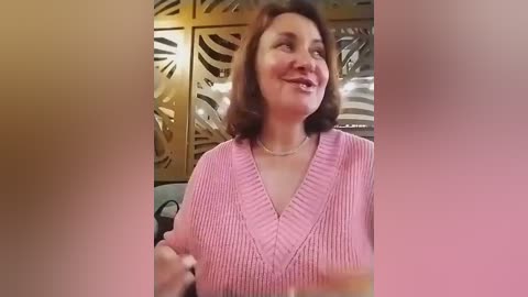 A video of a middle-aged woman with shoulder-length brown hair, wearing a pink V-neck sweater, smiling. Background features a golden, lattice-patterned wall and a blurred, warm-toned figure.