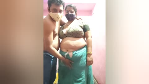 Video of a shirtless man wearing a surgical mask and a woman in a green sari with a polka-dotted bra, standing in a pink-walled room.