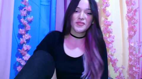 Video of a young woman with long, dark hair and a black choker, dressed in a black top, seated in front of a backdrop with pink and blue curtains adorned with floral patterns.