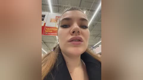 Video of a young woman with light skin, pigtails, and a nose piercing, looking upwards in a brightly lit supermarket.
