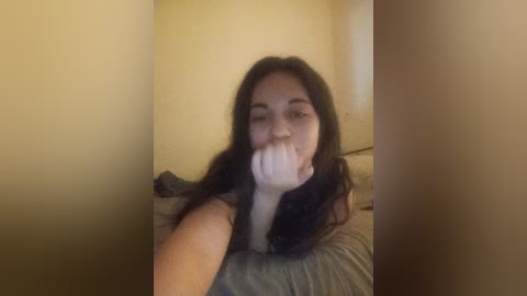 Video of a young woman with long black hair, fair skin, and a playful expression, making a bunny ear with her fingers, in a dimly lit room with yellow walls.