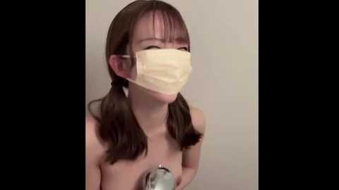 Video of a young Asian woman with long brown hair in pigtails, wearing a yellow surgical mask, holding a showerhead, topless, against a plain white background.