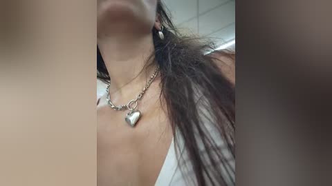 Video of a woman with long, dark hair, wearing a silver heart necklace, in a bathroom setting, blurred faces in the foreground and background.