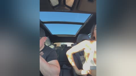 Video of a car interior with two men in the back seat; one with a white T-shirt, the other with a yellow shirt. The sunroof is open, revealing a clear blue sky.