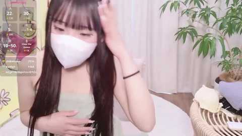 Video of an Asian woman with long black hair, wearing a white mask and green top, adjusting her hair. Background includes a plant and wicker basket.