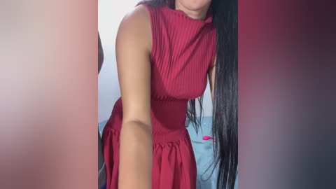 Video of a woman with long black hair, wearing a sleeveless, ribbed red dress, bending slightly, with blurred background.