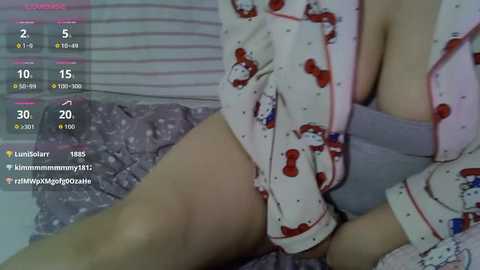 A video of a woman in a pink, Hello Kitty pajama top, revealing her large breasts and light skin, sitting on a bed with striped sheets.