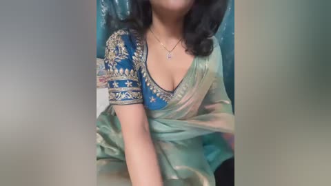 Video of a woman in a blue and green sari with intricate gold embroidery, wearing a necklace, seated against a blurred background.
