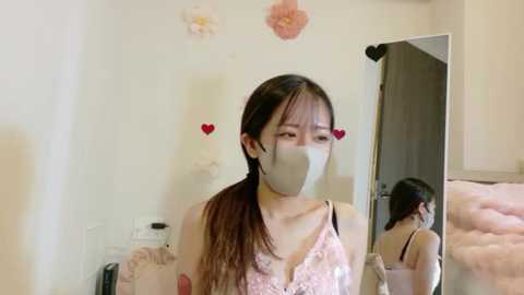 Media: Video of an East Asian woman with long dark hair, wearing a pink floral dress, face mask, and heart stickers, standing in a softly lit room with a mirror reflecting her profile.