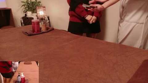 Video of a woman in a red sweater and black skirt taking a selfie in a dimly lit, cozy room with a brown quilted massage table, a tray of massage oils, and a lit candle.