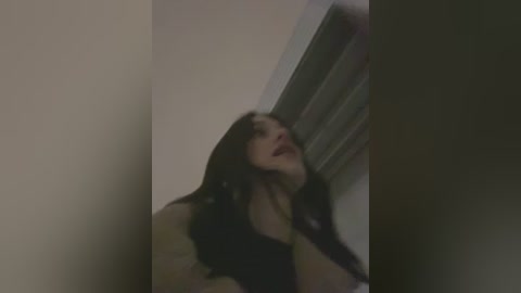 A blurry video of a woman with long dark hair, wearing a black top, leaning against a beige wall with a window partially visible.