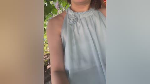 Video of a woman with light skin wearing a sleeveless, light blue blouse with a floral pattern, standing outdoors among greenery and blooming flowers.