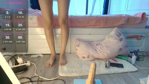 Video of a barefoot person standing on a rug in a messy bedroom with a bed, pillows, and scattered items including a vibrator and bottle.