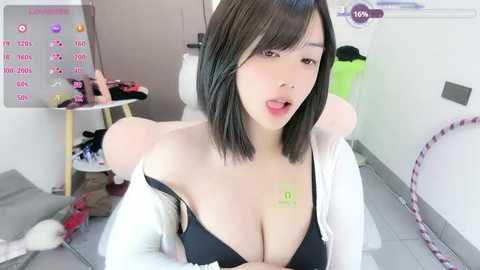 A video of an Asian woman with a slim figure, short black hair, and fair skin, wearing a black bra and off-shoulder white top. The background shows a cluttered laundry room with a washing machine and clothes scattered on the floor.