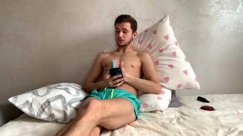 Media: Video of a young, shirtless, fair-skinned man with short brown hair, wearing green shorts, playing with a smartphone on a bed with white pillows and pink heart-patterned sheets.