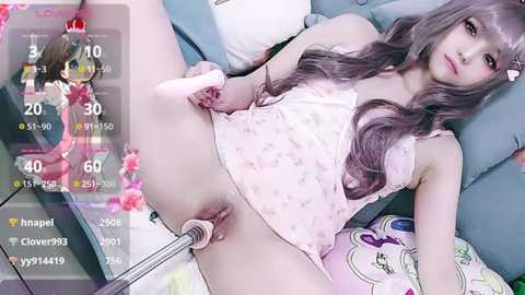 A video of an East Asian woman with long, wavy, gray hair, wearing a pink floral top, lying on a bed. A smartphone screen with a video recording feature is overlaid, showing a virtual character in a game.