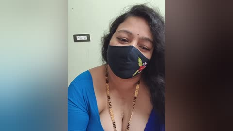 Video of a middle-aged South Asian woman with dark hair, wearing a blue blouse and black face mask with colorful embroidery, standing in a room with a beige wall and a light switch.