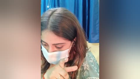 Media: Video of a young South Asian woman with light brown skin and long, wavy brown hair, wearing a light blue surgical mask, seated in a room with a blue curtain in the background.