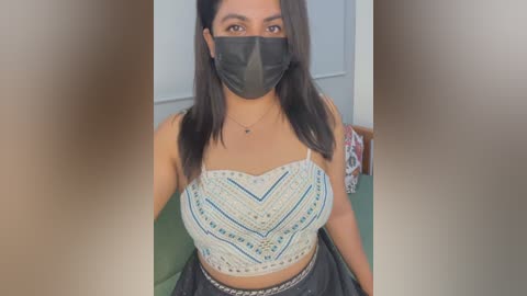 Video of a young woman with medium skin tone, wearing a black face mask, black tank top with white geometric patterns, and high-waisted black pants, sitting on a green couch in a brightly lit room.