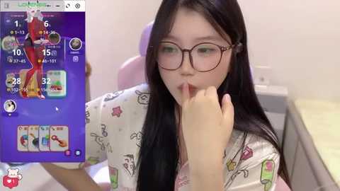 A video of an Asian woman with long black hair and glasses, wearing a pink shirt with cartoon characters, playing a video game on a smartphone.