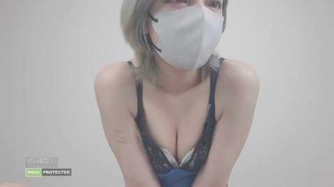 Video of a fair-skinned woman with shoulder-length, platinum blonde hair, wearing a white surgical mask and a blue lace bra, with visible cleavage, against a plain, light gray background.