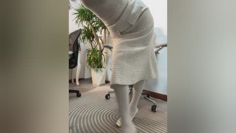 Media: Video of a woman in a beige dress, bending over a desk, with a large potted plant and a black office chair in the background.