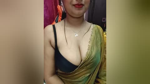 Video of a South Asian woman with fair skin, wearing a green and yellow sari, black bra, and necklace. Background features a colorful, patterned wall.