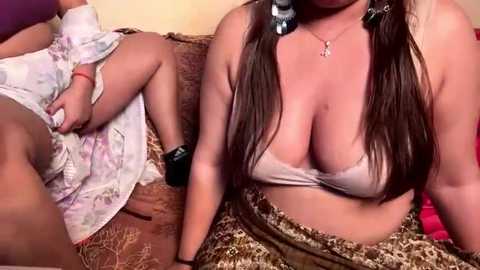 Video of a busty, light-skinned woman in a white bra, leopard-print skirt, and black wrist cuffs, sitting on a couch, while another woman, partially visible, lies on the floor in floral pajamas.