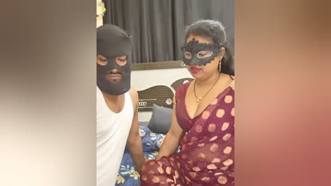 A video of two individuals in a bedroom, both wearing black masks and Indian traditional attire. The man is in a white tank top, the woman in a maroon sari with polka dots.