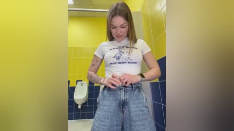 Video of a young woman with fair skin, long brown hair, and tattoos, wearing a white graphic tee and high-waisted blue jeans, adjusting her waistband in a yellow and blue-tiled bathroom with a urinal and mirror.