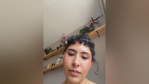 Media: Video of a young woman with short curly hair and light brown skin, wearing makeup, standing in a cozy room with wooden shelves displaying decorative items and a floral arrangement.