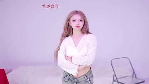 Video of a slender, fair-skinned, long-haired East Asian woman in a white, crocheted crop top and denim shorts, standing confidently with crossed arms. Background includes a plain white wall, a metal folding chair, and Chinese text.