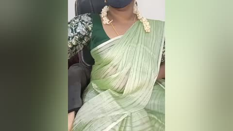 Video of a woman wearing a green saree with a floral blouse, black mask, and gold necklace, seated on a black chair.