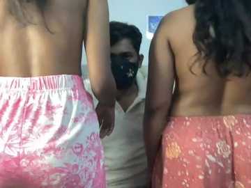 Video of two women in pink and red patterned skirts, topless, standing in front of a man wearing a mask, indoors.