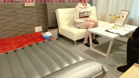 Video of a woman in a beige sweater and black shorts sitting on a white couch in a minimalist room with a grey carpet, a silver inflatable bed, and a white table with a golden trophy.