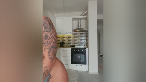 Media: A video of a tattooed man with a large, intricate sleeve tattoo, standing in a modern, white kitchen with stainless steel appliances and a backsplash.