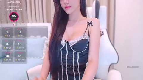 Video of an Asian woman with long black hair, wearing a black corset with white lace trim and a small bow, sitting on a white gaming chair in a minimalist room.