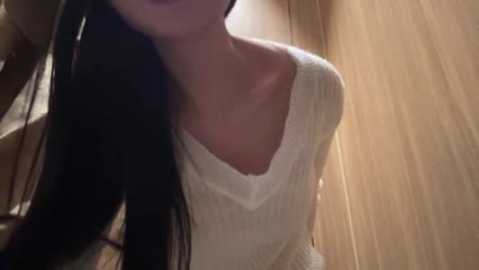 Media: Video of a young woman with long black hair, wearing a white, knitted sweater, standing against a wooden background. Her head is tilted slightly, and her lips are painted dark purple.