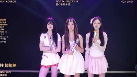 Video of three young Asian women on stage, wearing light pastel dresses, holding microphones, standing against a dark background.