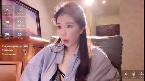 Video of a young Asian woman with long black hair, wearing a blue shirt, sitting on a couch in a dimly lit room with a TV displaying sports scores in the background.