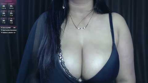Video of a woman with long black hair, wearing a black lace bra that reveals ample cleavage, against a dark background.