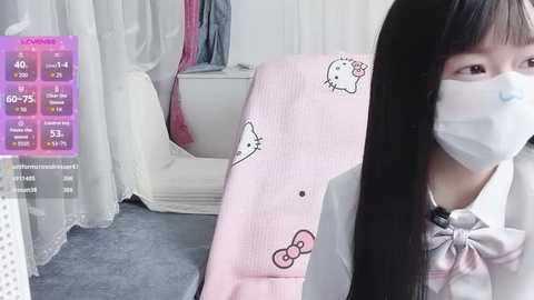 Video of an Asian woman with long black hair, wearing a white face mask, pink Hello Kitty blanket, and a white blouse, in a room with a bed and pink curtains.