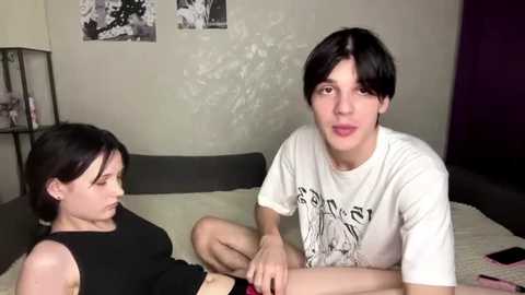 Video of a young, slender woman with short black hair, wearing a black tank top, sitting on a bed. A young man with straight black hair and pale skin, wearing a white Nirvana t-shirt, sits beside her. Background includes a plain wall with black and white photos.