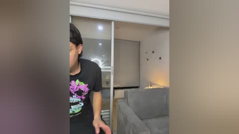 Video of a man with short black hair, wearing a black T-shirt with colorful floral graphics, standing in a modern living room with a gray sofa, white walls, and a glass sliding door.