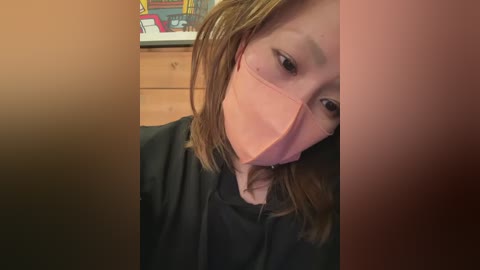 A video of a young woman with light skin, wearing a pink surgical mask, brown hair, and a black hoodie, standing indoors with a wooden dresser in the background.