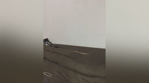 Video of a bare, white-walled room with a black, crumpled blanket on the floor, slightly to the left. A single white headphone cord is visible. The room appears empty and dimly lit.